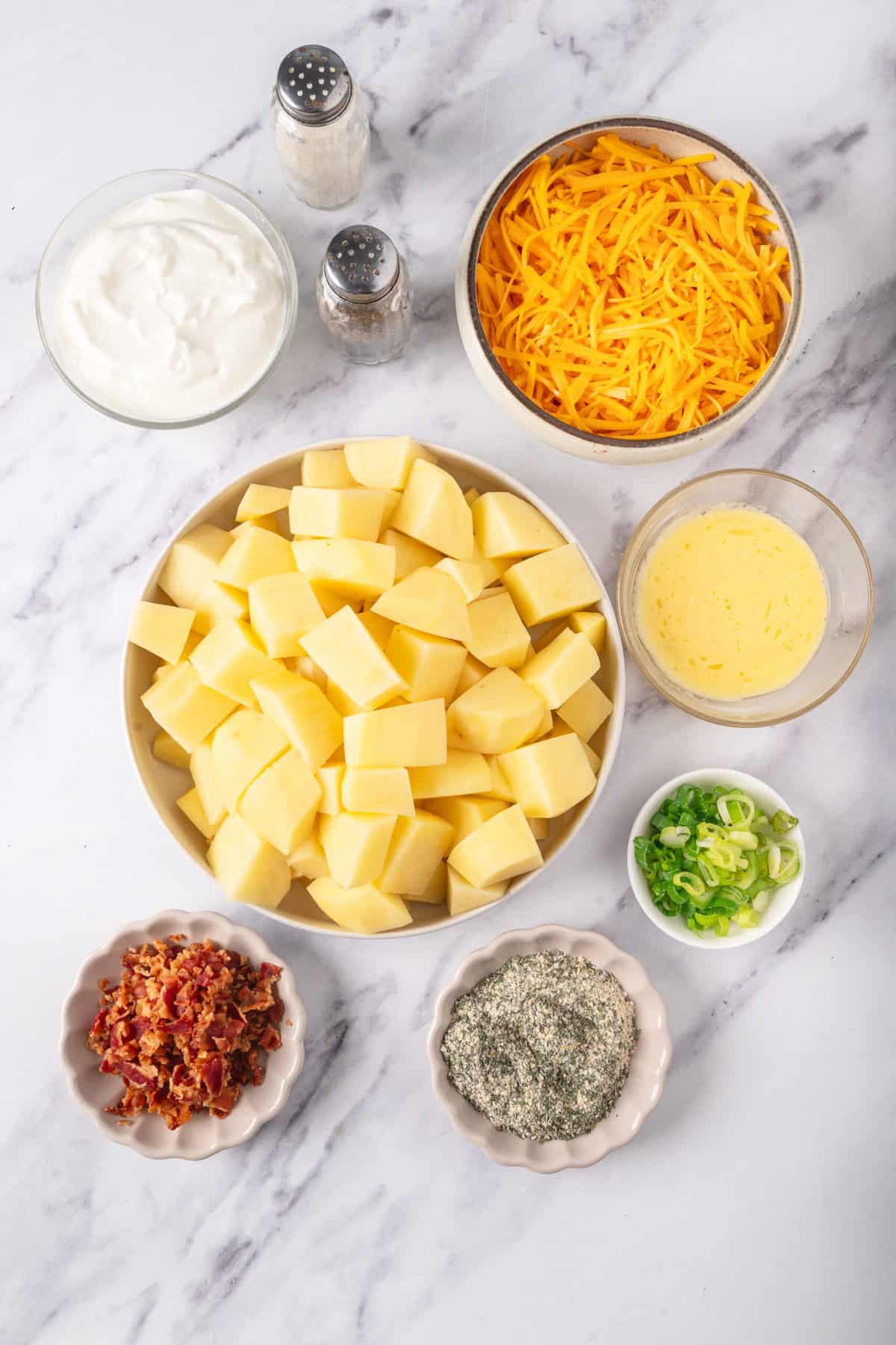 Creamy potato cheese casserole ingredients with shredded cheddar, chopped potatoes, sour cream, green onions, bacon bits, and seasonings on a marble surface.