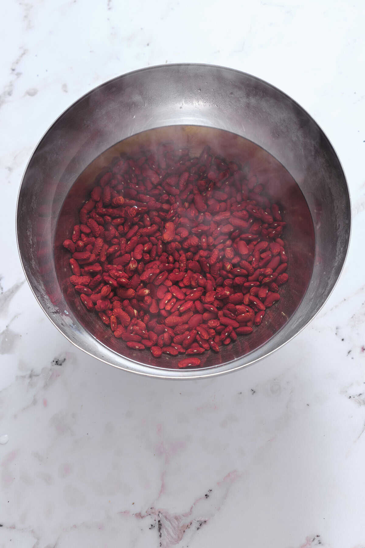 Steaming red adzuki beans in a stainless steel bowl on a white marble surface.