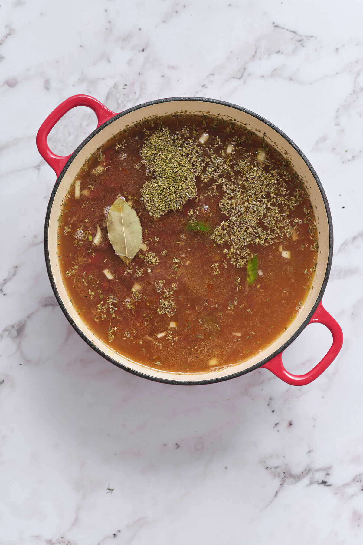 Herbal and vegetable soup in a red pot, featuring bay leaves and herbs, on a white marble countertop. Perfect for healthy eating and nutritious recipes.