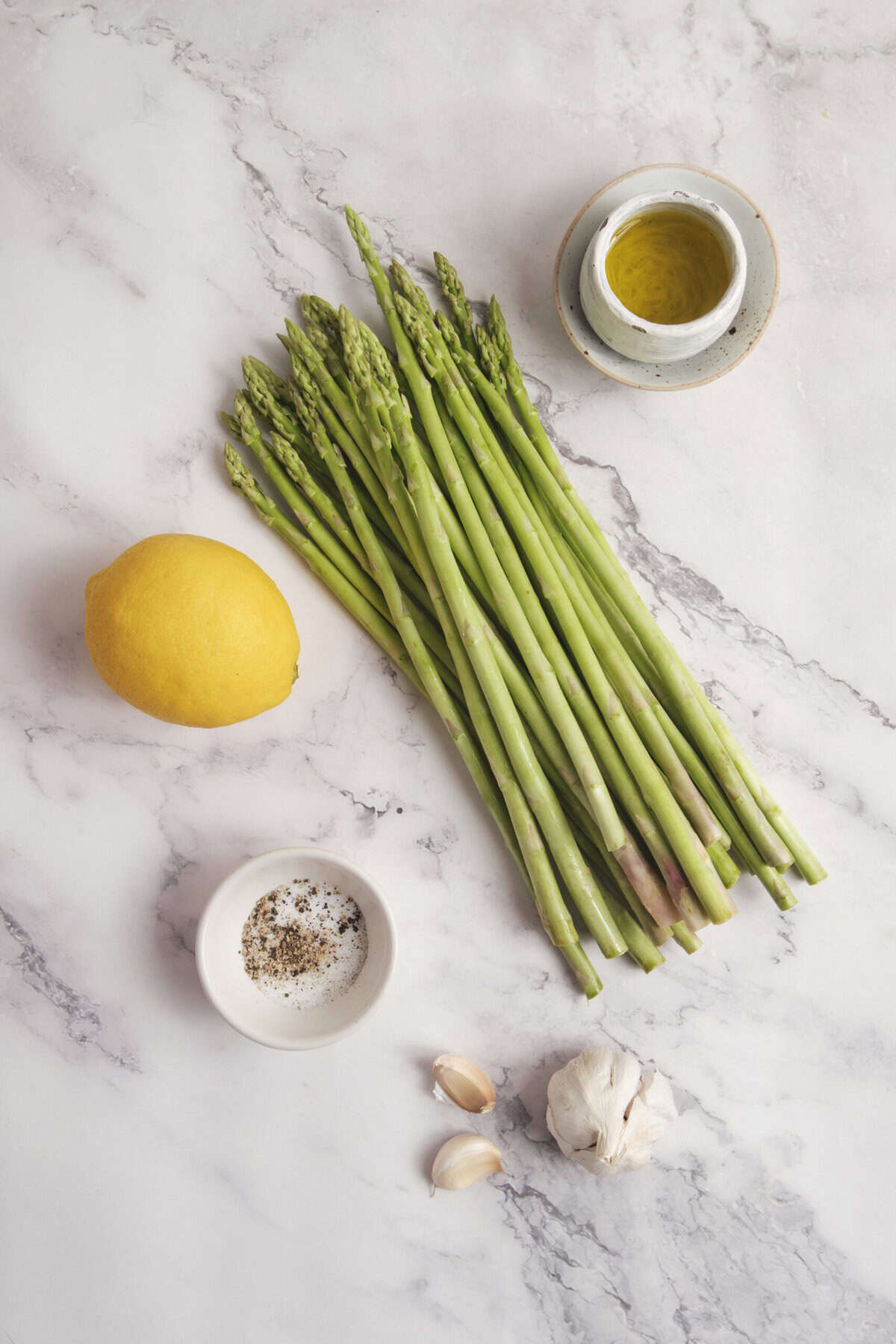 Fresh asparagus with lemon, garlic, sea salt, black pepper, and olive oil on a white marble surface, perfect for healthy recipes and clean eating.