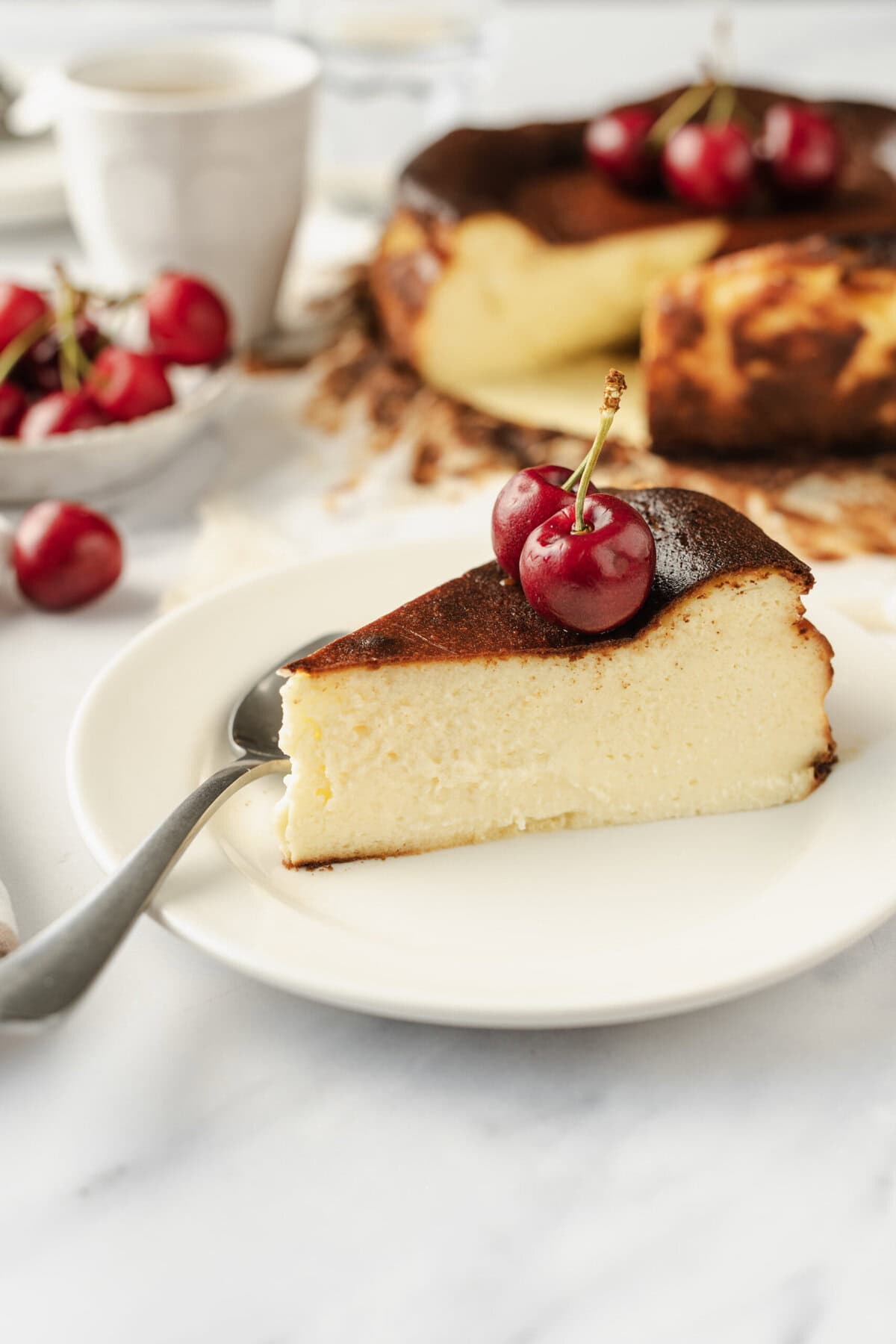 Cream cheese cheesecake topped with fresh cherries on a white plate with a silver fork, showcasing a delicious and healthy dessert recipe from Food Faith Fitness.