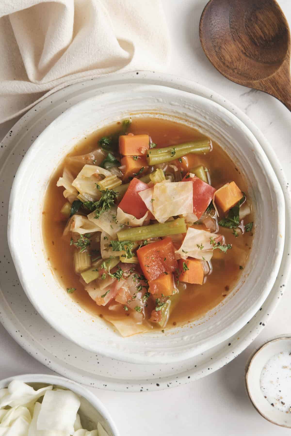 Vegan vegetable soup with fresh carrots, celery, cabbage and tomatoes in clear broth, served in a white bowl on a rustic background.