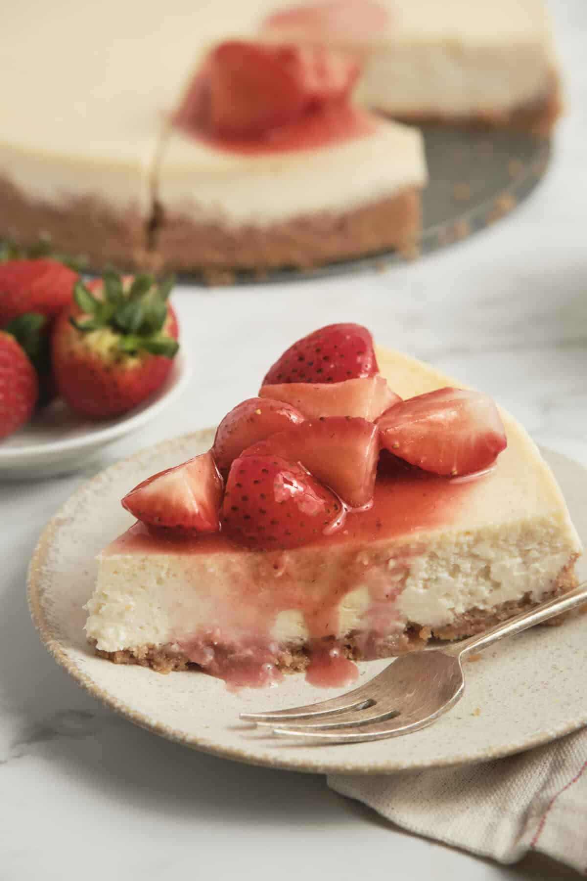 Fresh strawberry cheesecake topped with strawberry glaze, served on a white plate with whole strawberries and a sliced cheesecake in the background. Perfect for healthy dessert recipes on Food Faith Fitness.