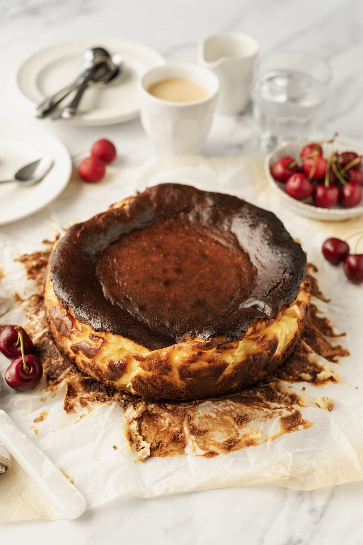 Rich chocolate cheesecake with a crispy crust, served on parchment paper with fresh cherries and a breakfast setting featuring coffee, milk, and cutlery. Perfect for a healthy dessert or indulgent treat.