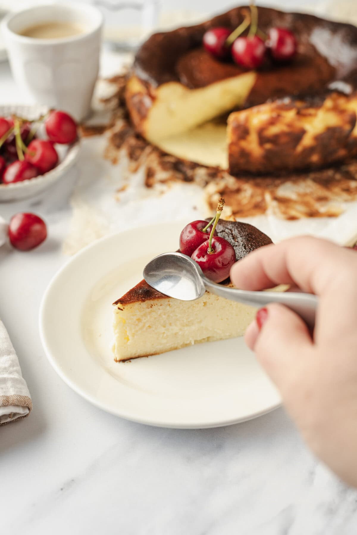 Creamy New York-style cheesecake topped with fresh cherries on a white plate, with a chocolate cherry cake and a coffee cup in the background, perfect for healthy dessert recipes.