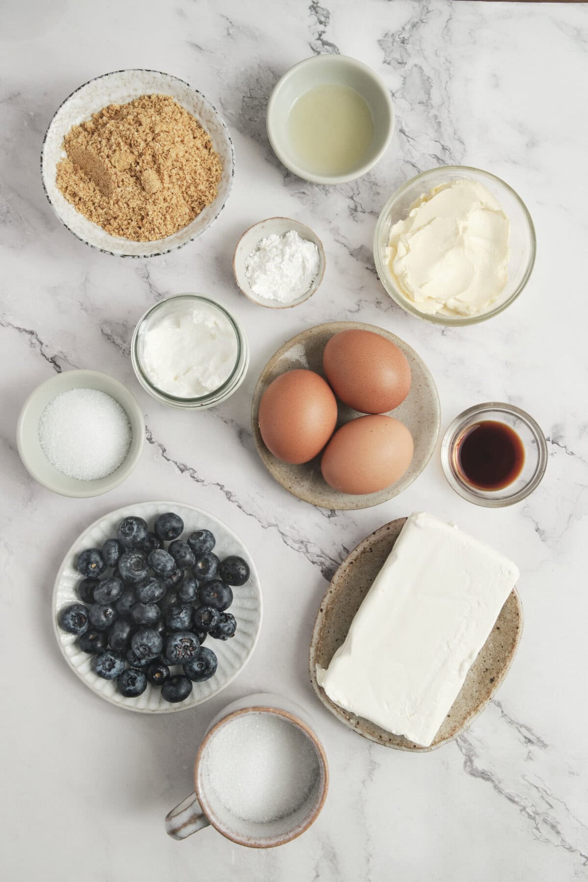 Flour, sugar, eggs, butter, blueberries, and vanilla extract on a marble surface, ingredients for a healthy baking recipe from Food Faith Fitness.