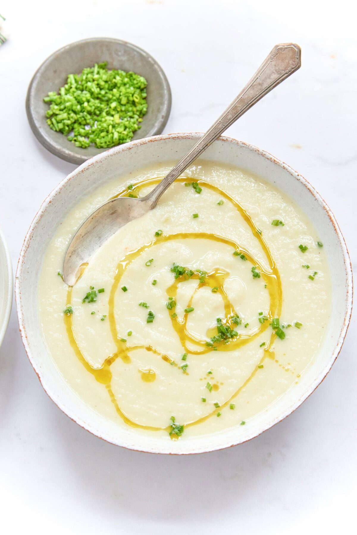 Creamy cauliflower soup garnished with chives and drizzled with olive oil, served in a white bowl. A small dish of chopped green onions is visible in the background. Perfect for healthy eating and nutritious recipes.