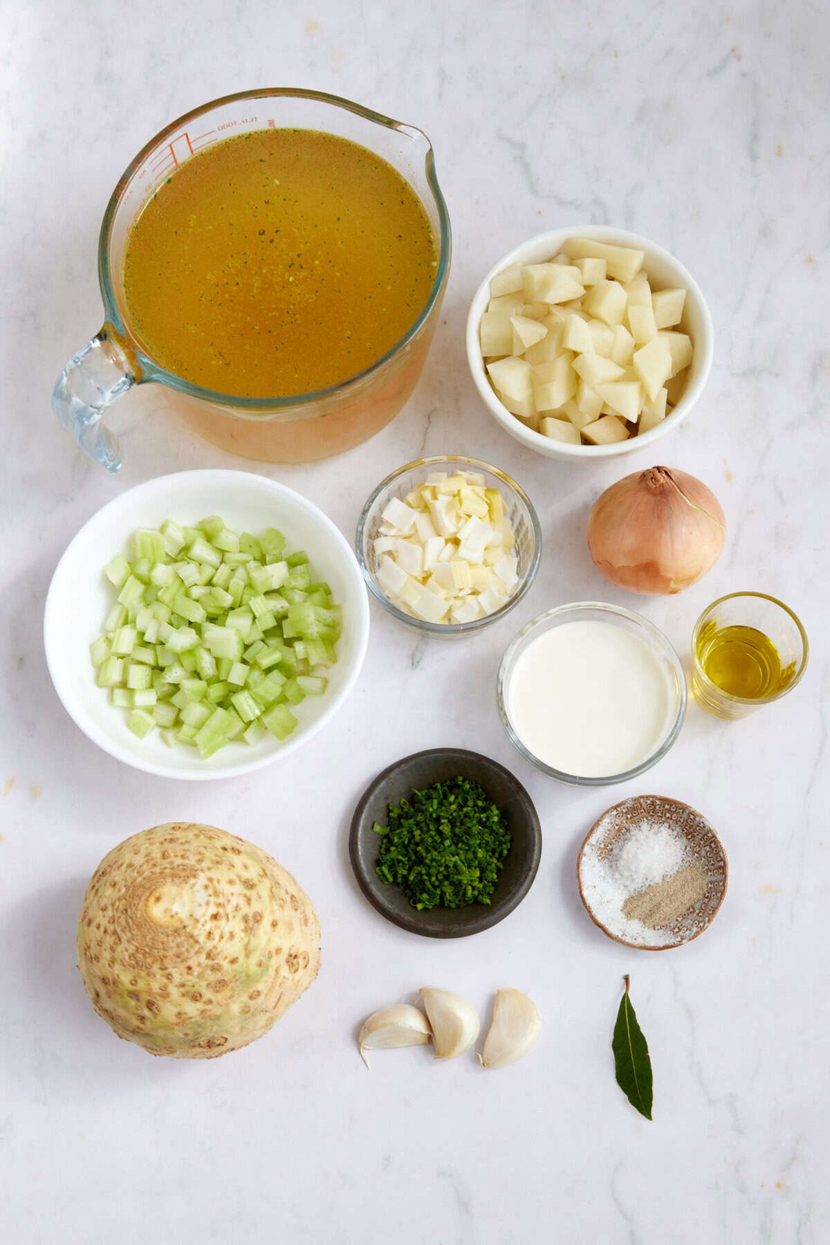 Vegetable soup ingredients including celery, onion, garlic, potatoes, vegetable broth, cream, and herbs arranged on a white surface.