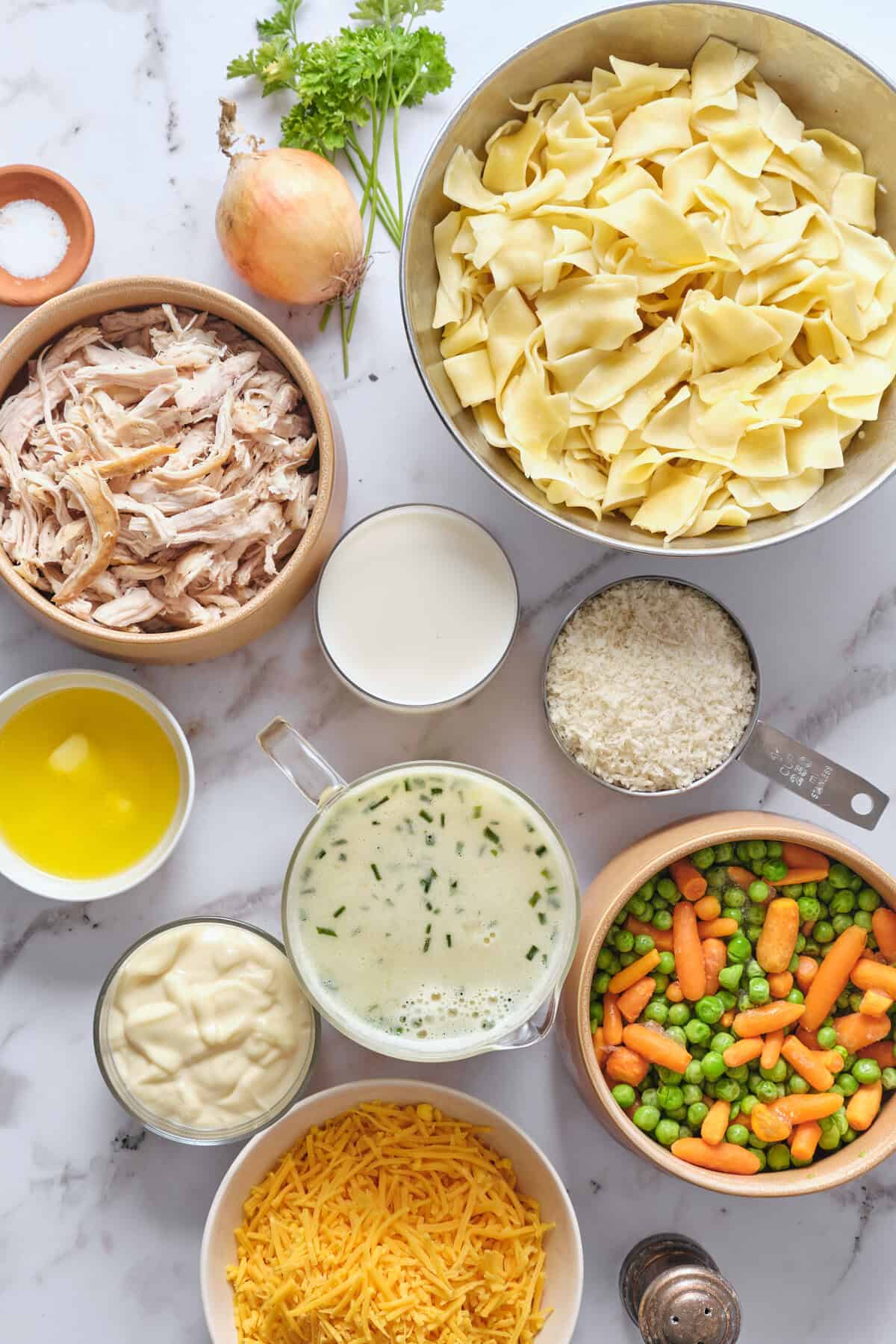 Shredded chicken, pasta, and vegetables for a healthy chicken alfredo recipe in bowls, with milk, cheese, and herbs on a marble countertop. Perfect for a nutritious dinner or meal prep.