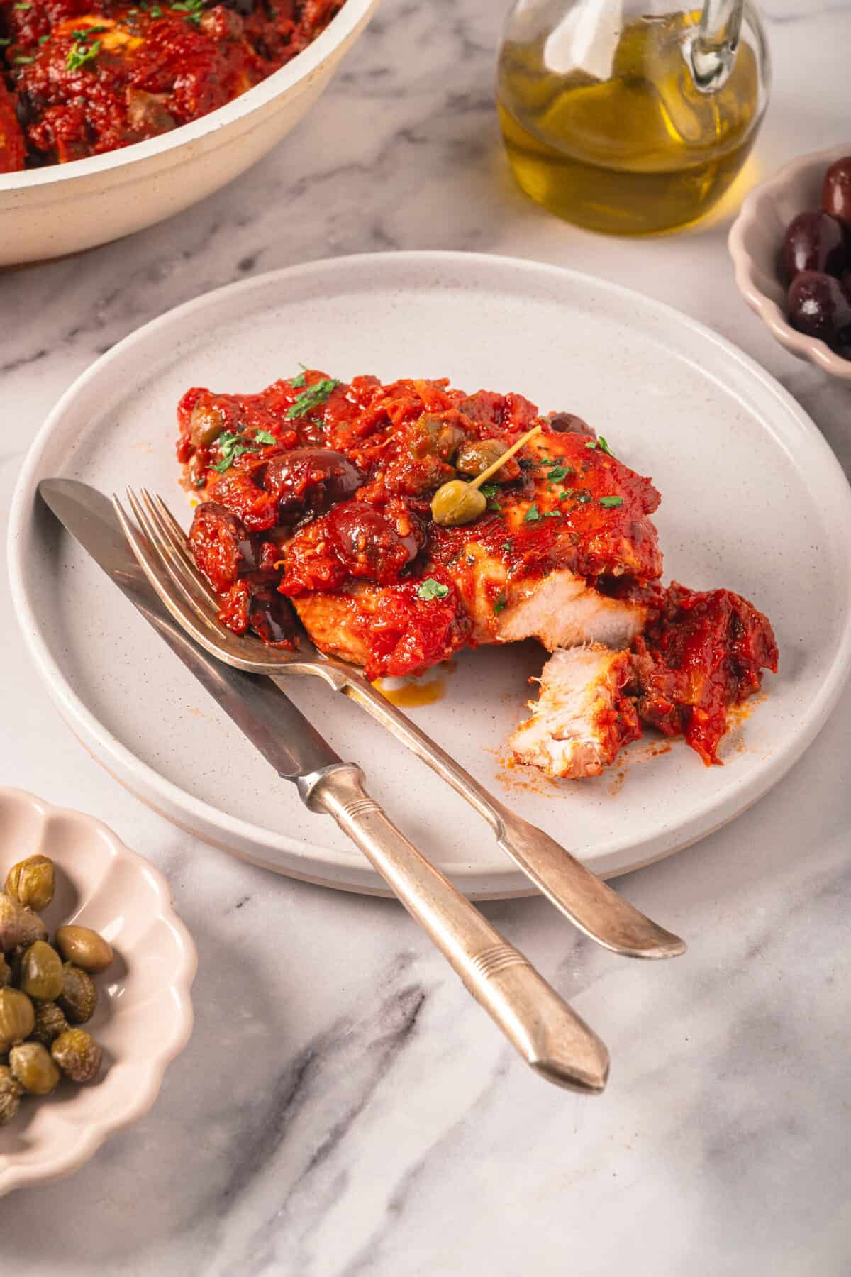 Hearty baked chicken topped with flavorful tomato sauce, olives, and herbs, served on a white plate with vintage silverware, surrounded by bowls of olives and olive oil on a marble countertop.