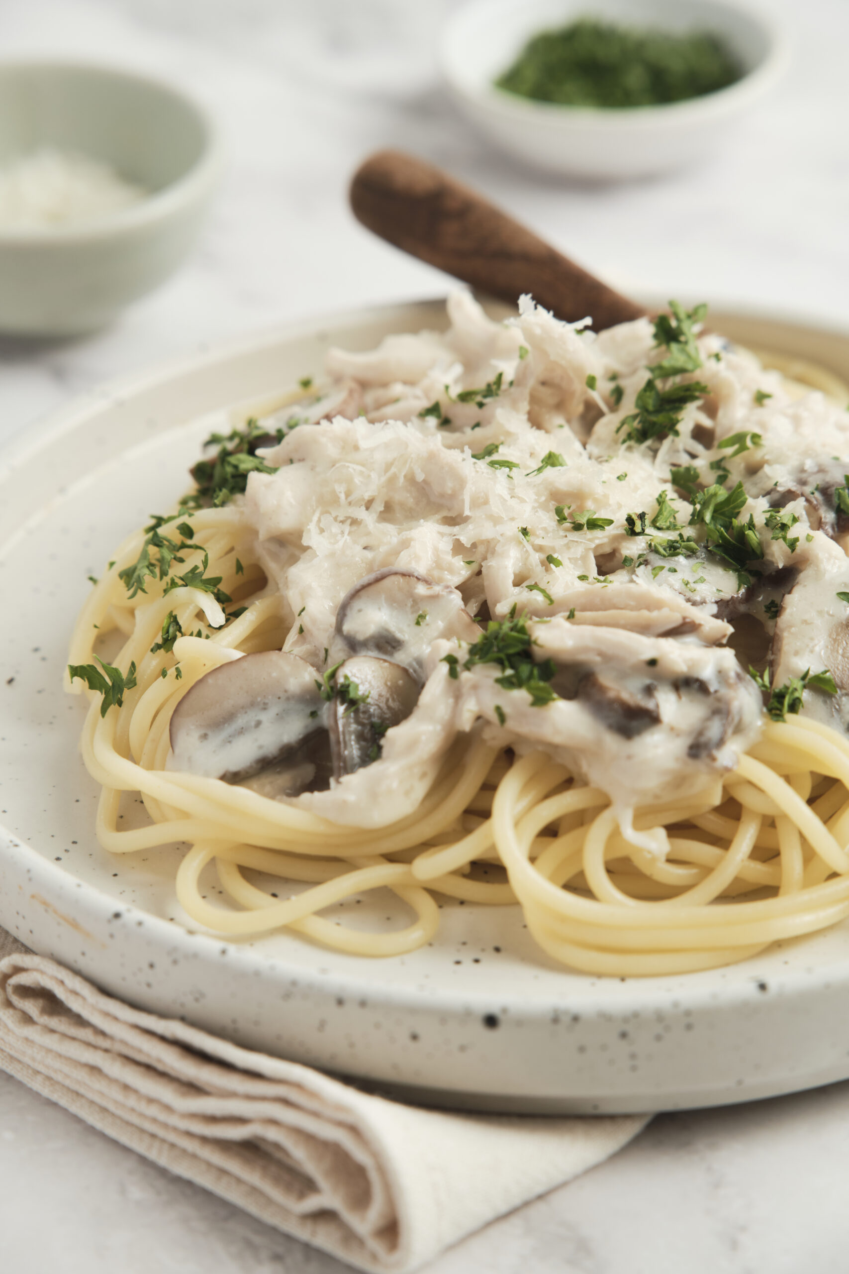 Creamy chicken and mushroom pasta dish topped with fresh parsley, served on a rustic speckled plate with a beige napkin and side bowls of herbs and grated cheese.