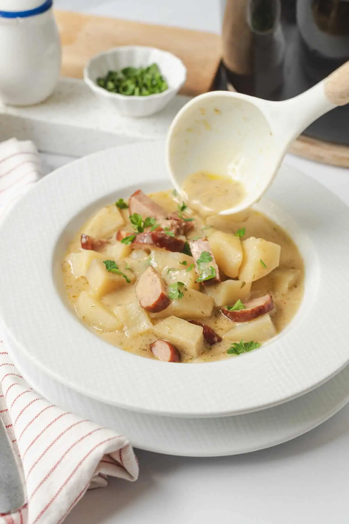 Creamy potato and bacon soup garnished with fresh herbs, served in a white bowl on a kitchen table. A ladle is pouring the soup, with green onions and kitchen utensils in the background. Perfect for a healthy, comfort food meal.