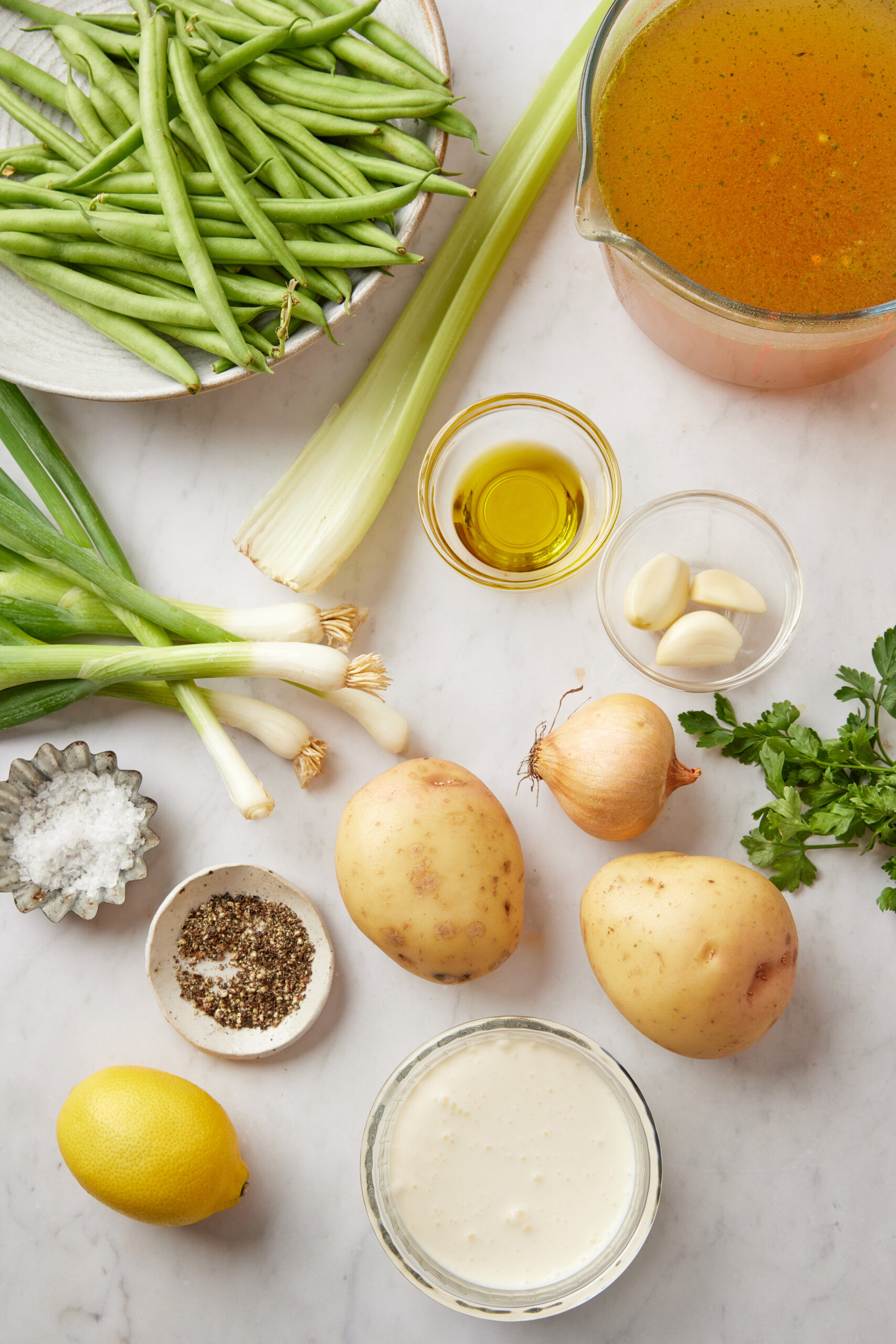 Fresh green beans, leeks, garlic, potatoes, onion, lemon, herbs, and vegetable broth ingredients on a white surface, ideal for healthy soup recipes and plant-based cooking, from Food Faith Fitness.