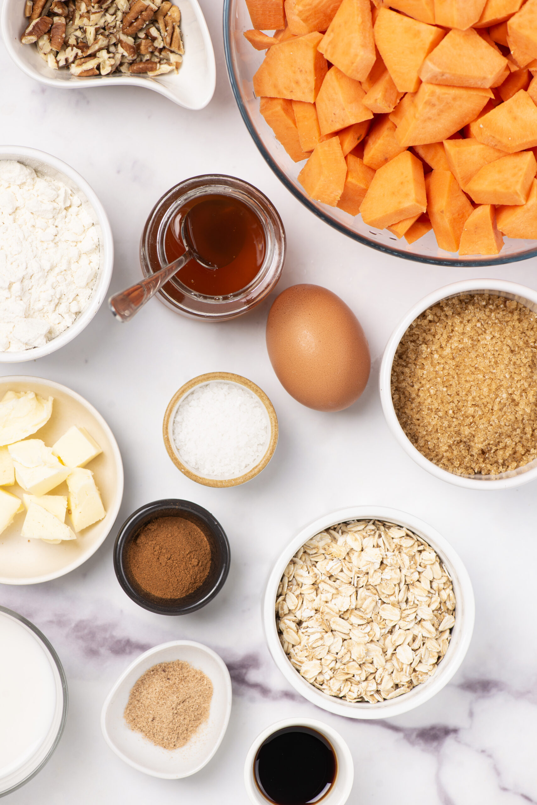 Sweet potato cubes, oats, eggs, butter, brown sugar, maple syrup, chopped pecans, vanilla extract, cinnamon, and flour on a white marble surface, ingredients for healthy breakfast recipes or protein-rich snacks.