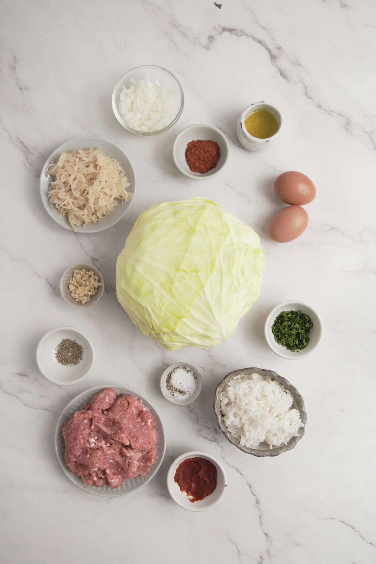 Fresh ingredients for healthy cabbage rolls, including ground meat, rice, cabbage, spices, eggs, and herbs, arranged on a white marble surface.