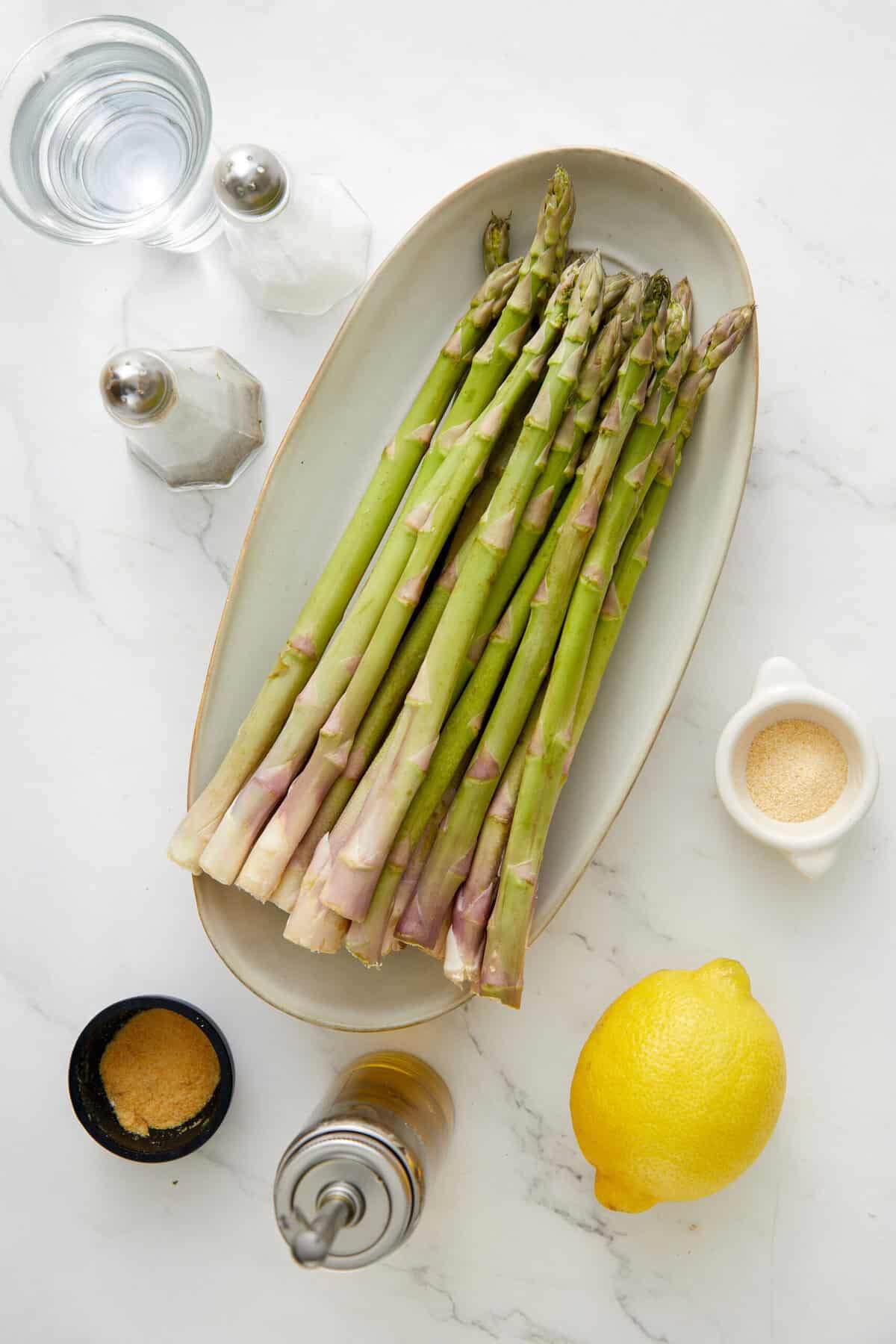 Fresh asparagus on a white platter with lemon, salt, and pepper shakers, ready for healthy meal preparation.