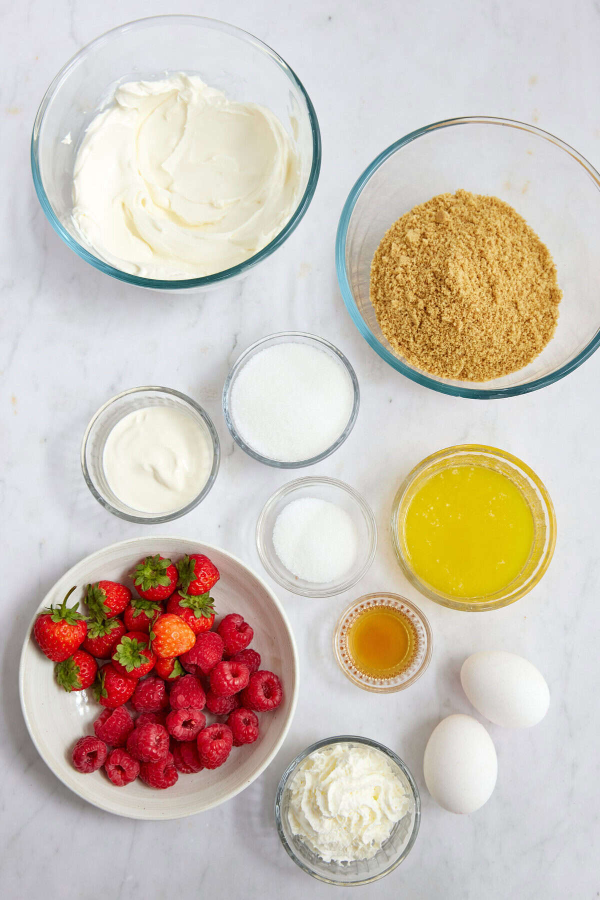 Fresh berries, cream cheese, and ingredients for healthy berry cheesecake on a white marble surface.