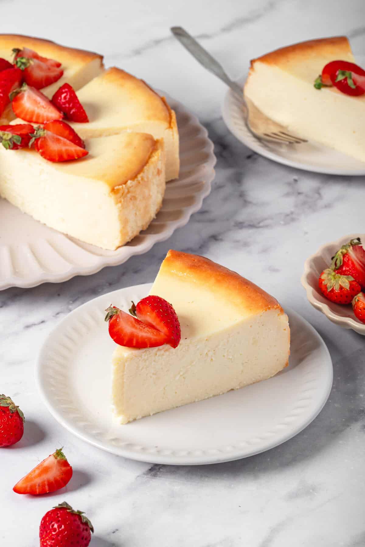 Creamy cheesecake topped with fresh strawberries on white plates, garnished with strawberry slices, served on a marble surface, highlighting healthy dessert recipes from Food Faith Fitness.