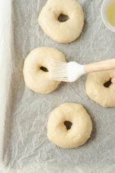 Brushing raw sourdough bagels with egg wash on a parchment-lined baking sheet.