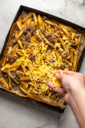 Sprinkling shredded cheddar cheese over cheeseburger fries on a baking sheet.