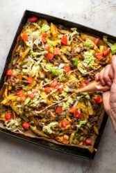 Drizzling burger sauce over a baking sheet of Cheeseburger Fries with beef, cheese, lettuce, tomatoes, and pickles.