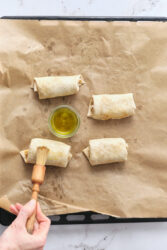 Brushing chimichangas with vegetable oil on a parchment-lined baking sheet.