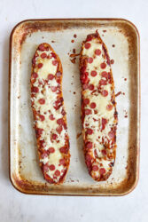 Two baked pizza bread halves with pepperoni and melted cheese on a baking sheet.