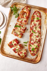Sliced pizza bread with pepperoni and basil on a baking sheet.